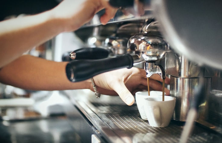 Closeup of unrecognizable caucasian barista making couple of espressos. He's placing cups under portafilter and pressing start button with his other hand. It's state of the art stainless steel espresso machine. Side view.