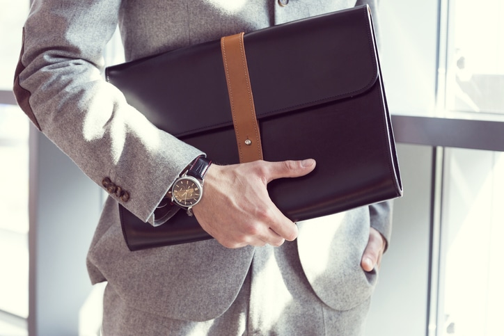 Businessman wearing grey suit standing in an office and holding black leather briefcase. Close up of hands, unrecognizable person.