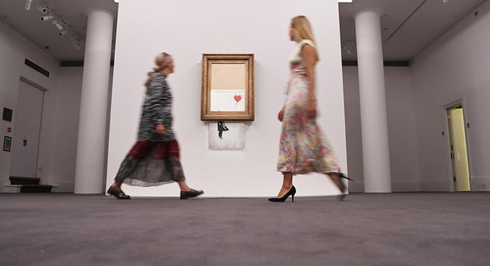 epa09445071 Gallery employees pose for photographers next to a painting entitled 'Love is in the Bin' by anonymous British street artist Banksy at Sotheby's auction house in London, Britain 03 September 2021. The painting was famously shredded by the artist at a Sotheby's auction in 2018 and it is expected to fetch GBP 4-6 million when it goes on sale on 14 October 2021.  EPA/FACUNDO ARRIZABALAGA